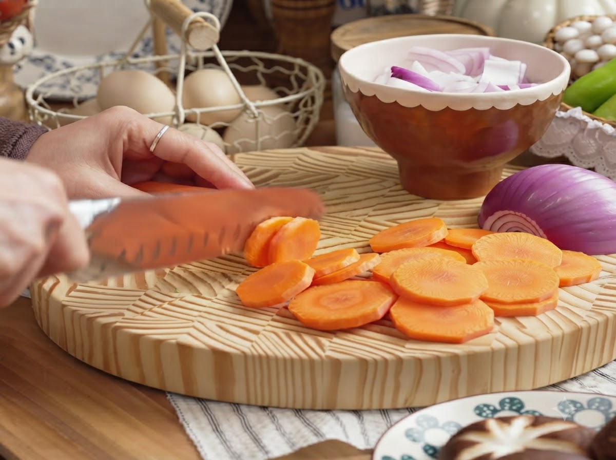 木製のまな板の上で、皮をむいたにんじんを輪切りにスライスしている手。