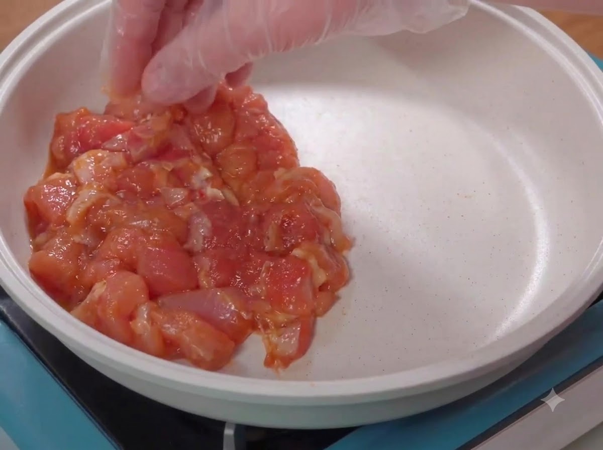 手袋をした手が、下味をつけた生の鶏肉を白いフライパンに並べている様子。