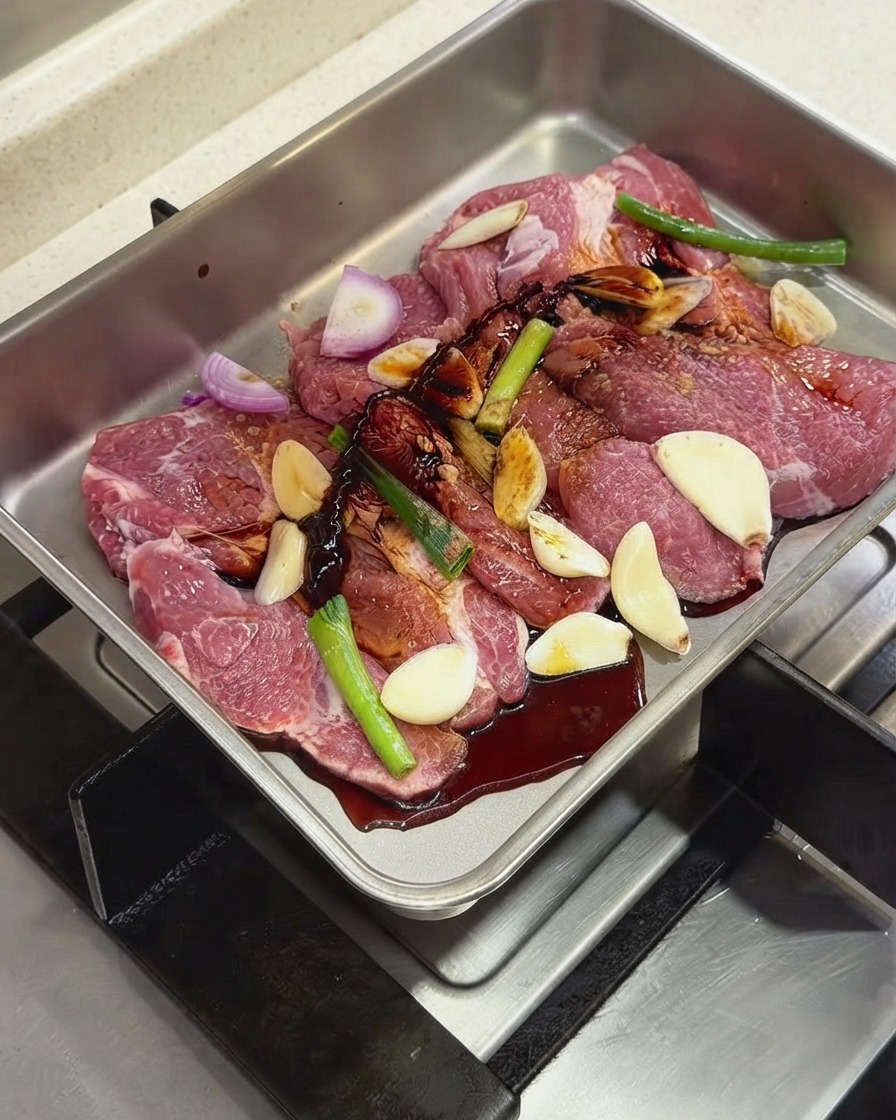 金属製トレイの中の豚肉と香味野菜に、濃い色のソースや調味料を注いでいる様子。