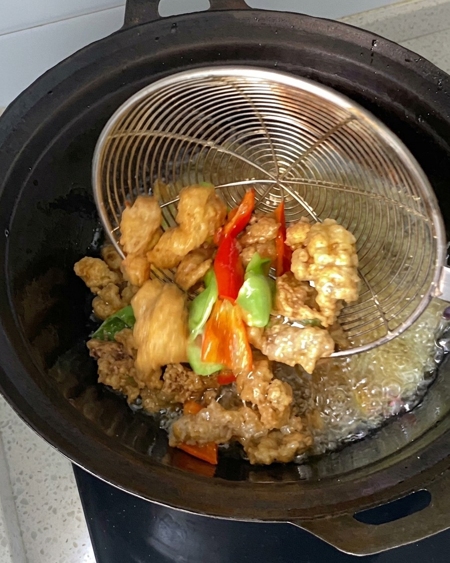 熱した油の中華鍋の中で、ワイヤーの strainer（網）を使ってマリネした肉とピーマンの断片がすくい上げられ、または入れられている様子。