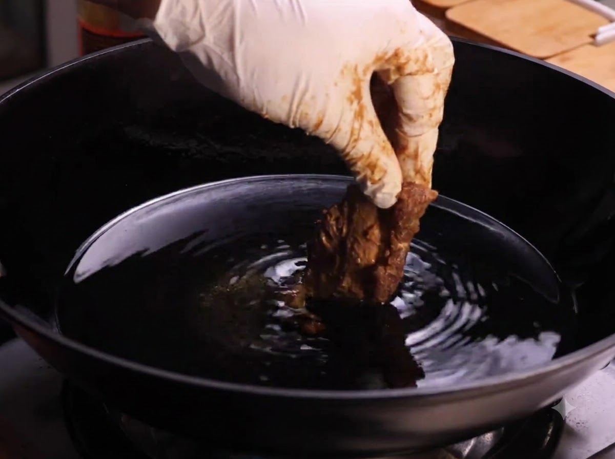 熱した油が入った中華鍋に、衣をつけた豚スペアリブの1切れを手袋をした手でそっと入れているところ。