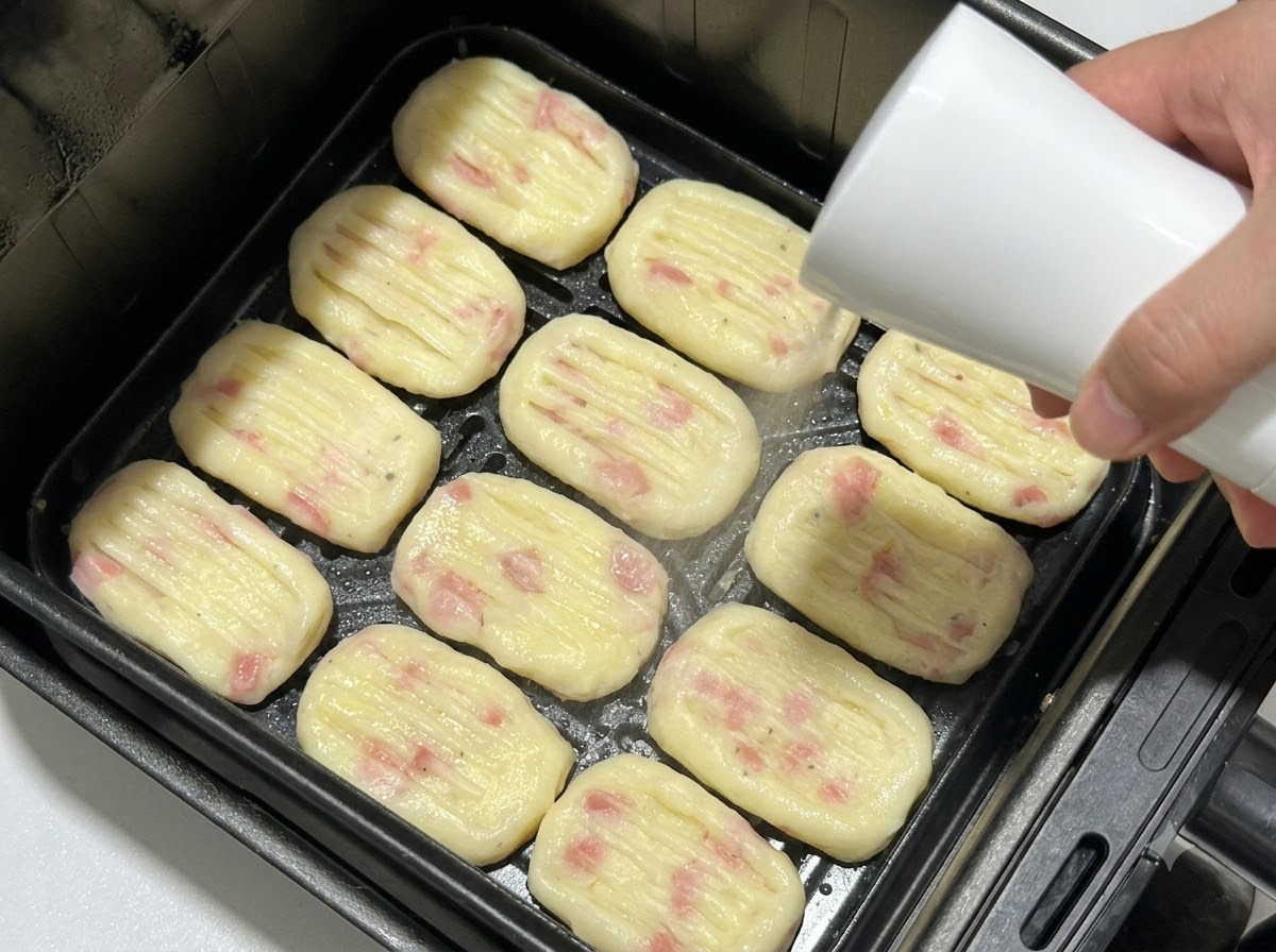 エアフライヤーのバスケットに並べられた成形済みのポテトパティに、スプレーボトルで油を吹きかけている様子。