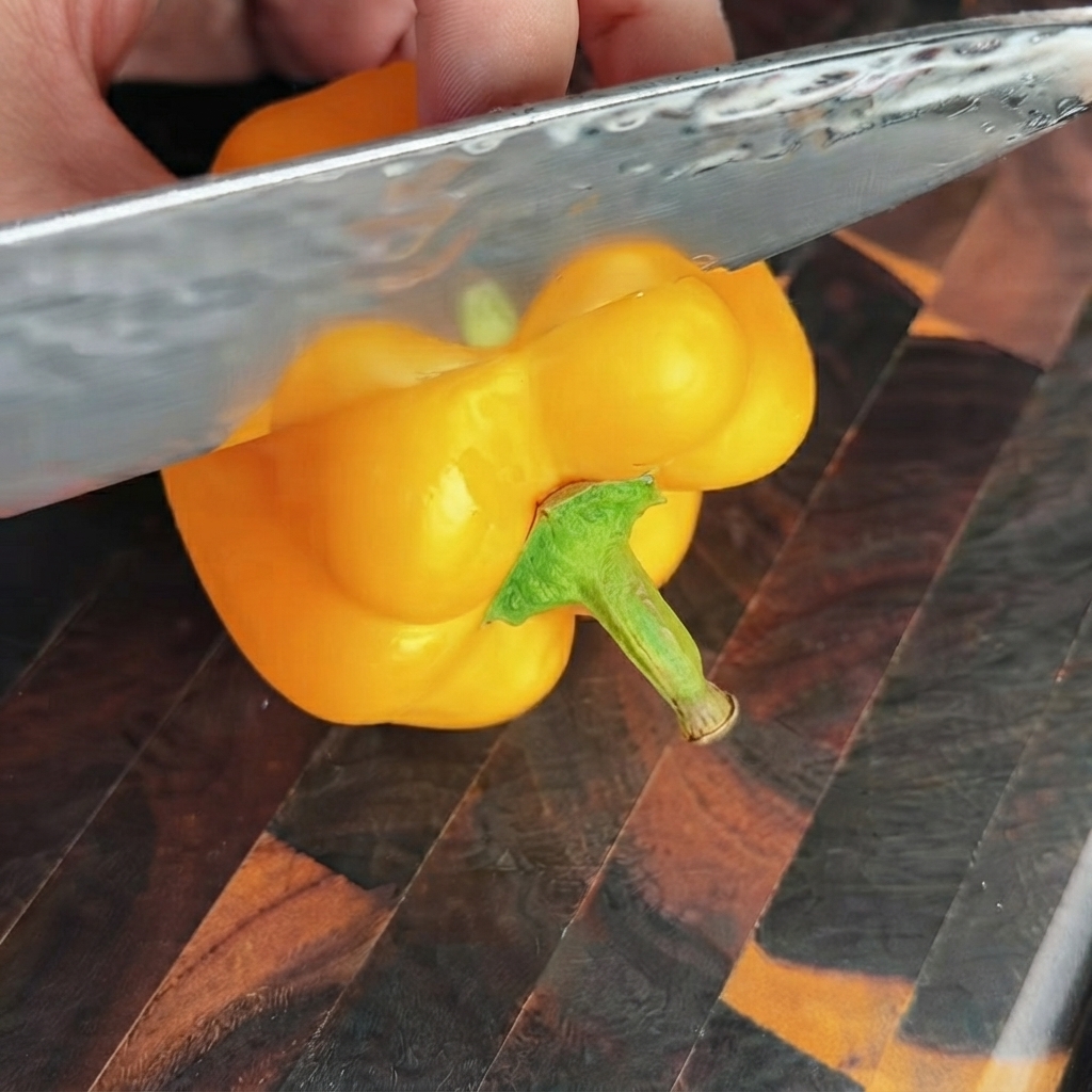 木製のまな板の上で、鋭い包丁を使って明るい黄色のパプリカの上部を切り落としている様子。