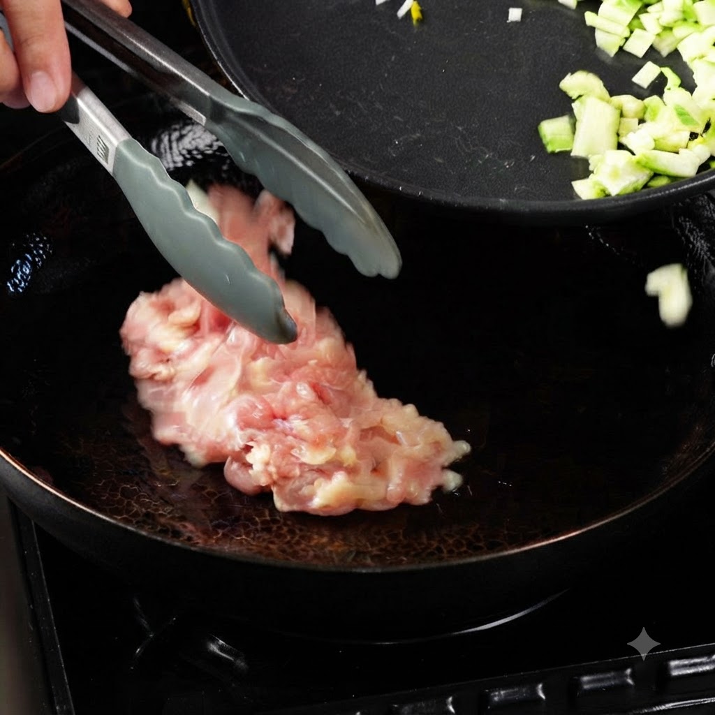 熱した油入りの中華鍋にトングで生の鶏肉片を入れているところ。隣には刻んだ野菜の入ったフライパンがある。