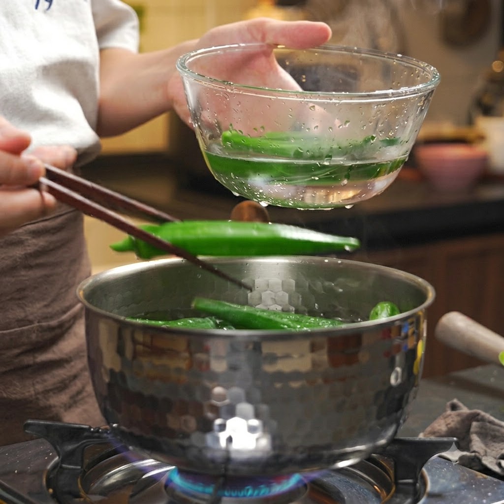 熱い鍋から箸を使って、茹で上がったオクラを氷水の入った透明なボウルに移している様子。