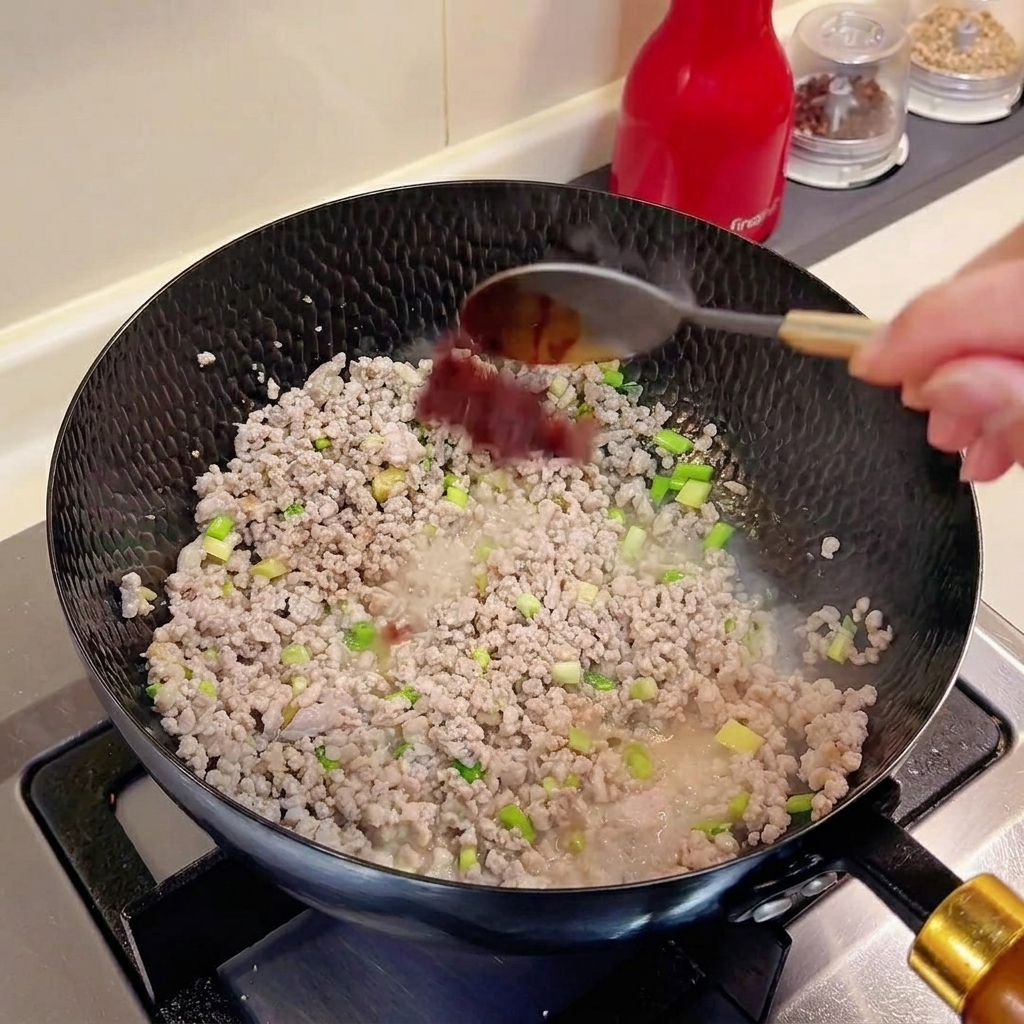 ひき肉と香味野菜の入った中華鍋にスプーン一杯の豆板醤が加えられている様子。