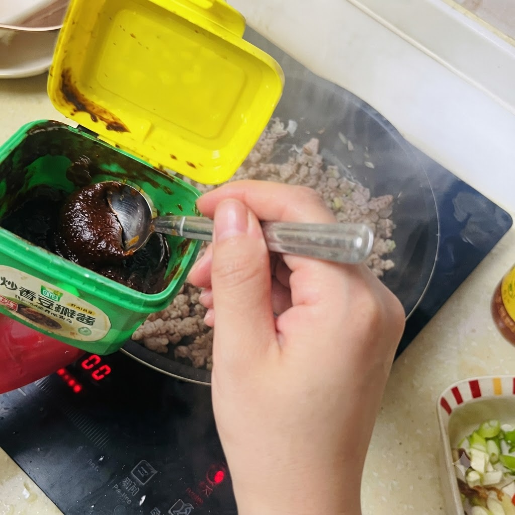 緑色の容器からスプーンですくった濃い色の味噌を炒めたひき肉に加えている様子。