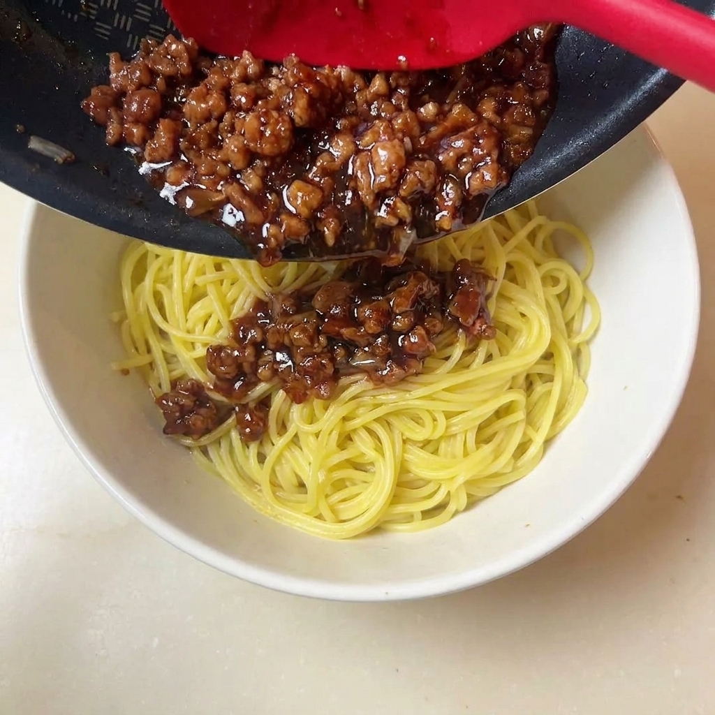 赤いスパチュラで濃厚な色の挽き肉ソースを黄色の麺の上にすくい入れている様子。