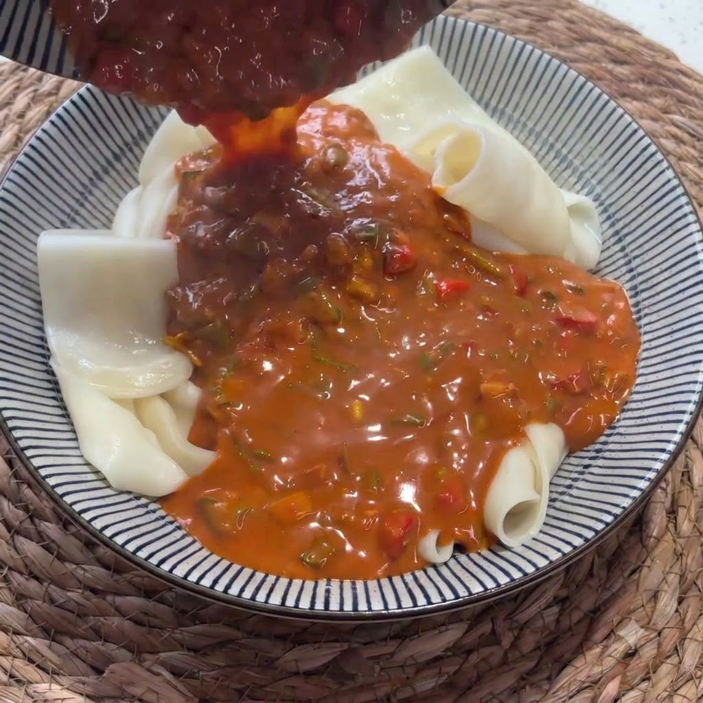 折り畳まれた幅広麺が入ったボウルに、おたまから濃厚な赤いソースが注がれている様子。