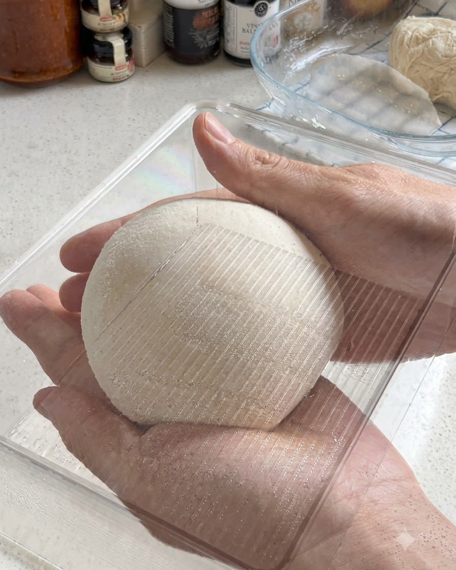 完璧に丸められたピザ生地のボールを、透明なプラスチック製の発酵容器に入れている2つの手。