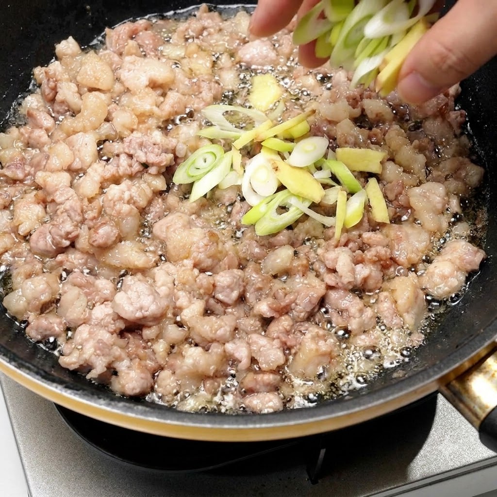 きつね色に炒められた豚肉が入ったフライパンに、刻みネギと生姜を直接加えている様子。