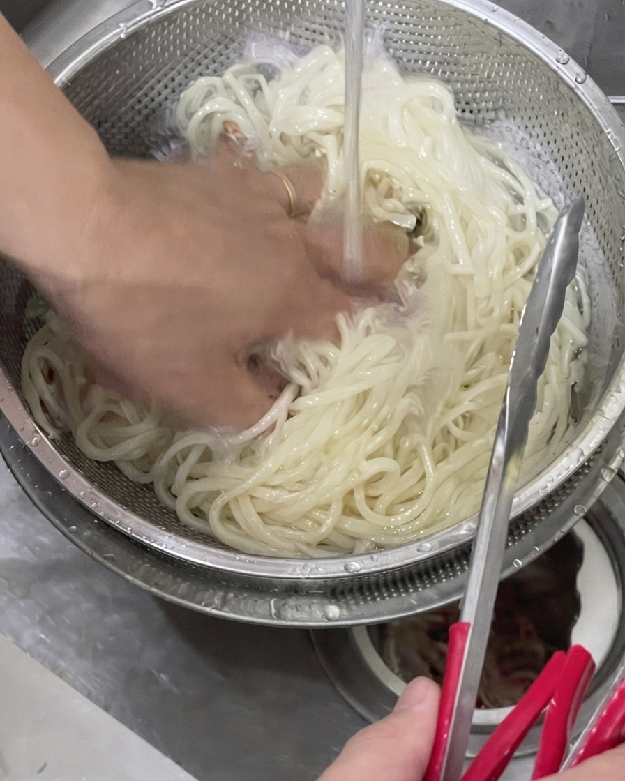 冷たい水道水の流れる金属製のザルの中で、手でうどんをほぐして洗っている様子。