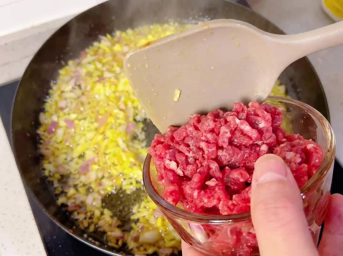 小さなガラスボウルから、炒めた玉ねぎとにんにくが入った熱々のフライパンに牛肉の角切りを入れているところ。