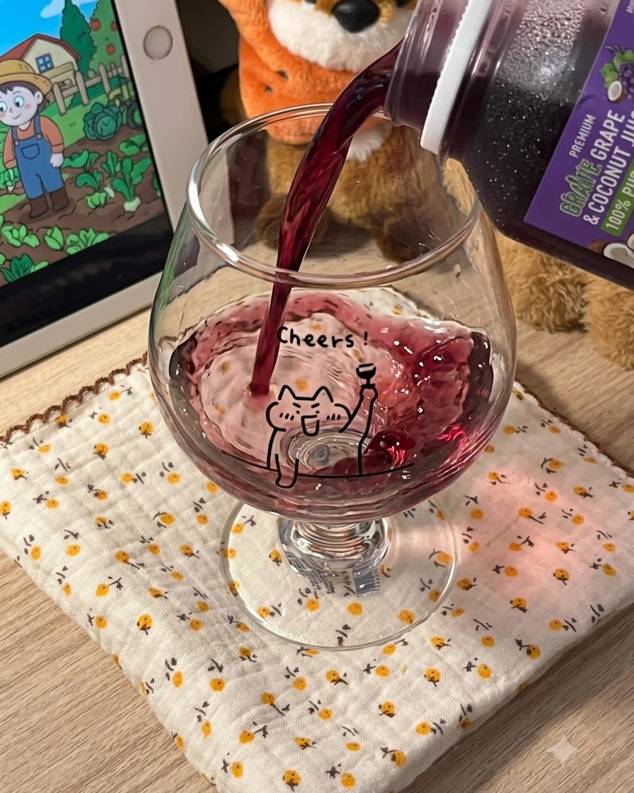 黄色い花柄のマットの上に置かれた、猫のイラスト付きの透明な足つきグラスに、濃い紫色のグレープジュースを注いでいる様子。