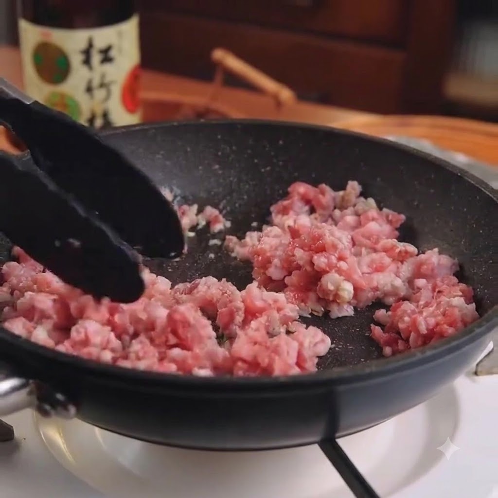 黒いフライパンの中で、調理用トングを使って生のエビひき肉とニンニクを炒めている様子。