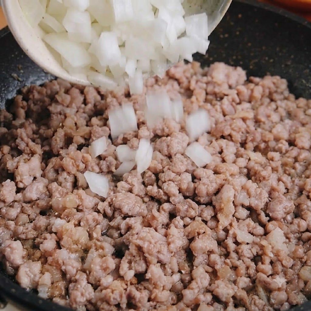 お椀から、炒めた豚ひき肉の入ったフライパンにダイス状の白玉ねぎを入れている様子。