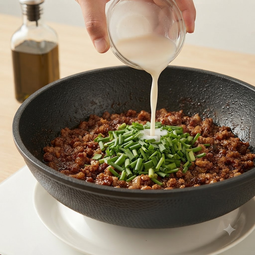 小さなガラス鉢から、ひき肉とニラの入ったフライパンに白い水溶き片栗粉を注いでいる手。