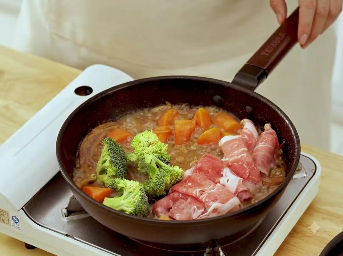 調理中の丸めた牛肉の隣で、新鮮な緑色のブロッコリーの小房を煮込み中のフライパンに加えている様子。