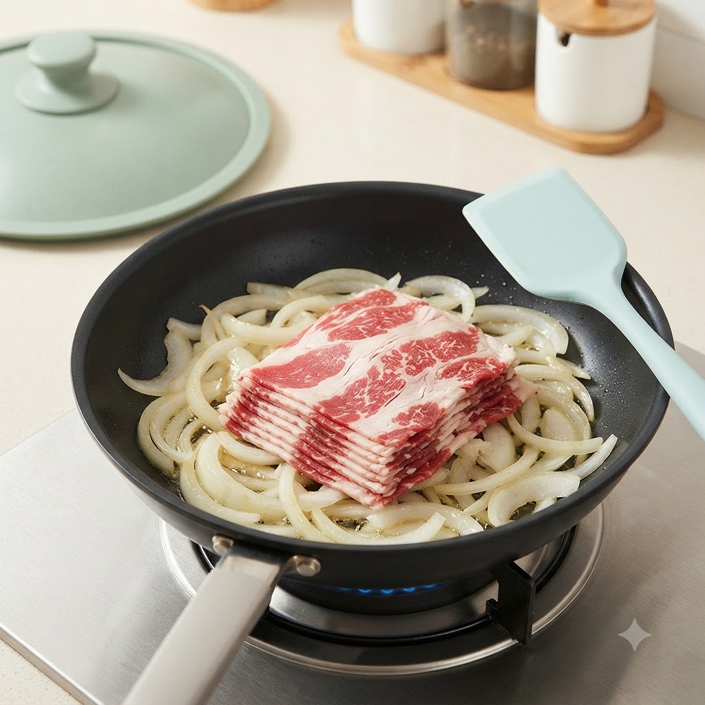 コンロに置かれたフライパンの中で、炒めた玉ねぎの上に牛肉の薄切りを並べている様子。