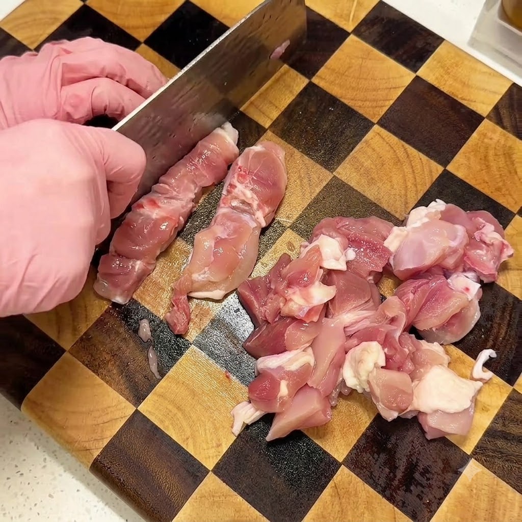 チェック柄の木製まな板の上で、ピンク色の手袋をはせた手が包丁を使って骨なしの鶏もも肉を切っている様子。