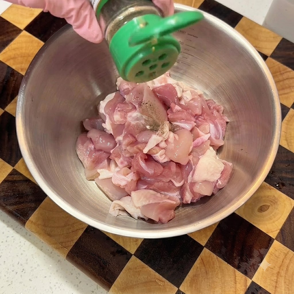 金属製のボウルに入った切った鶏肉に、緑色のシェーカーから黒こしょうを振っている様子。