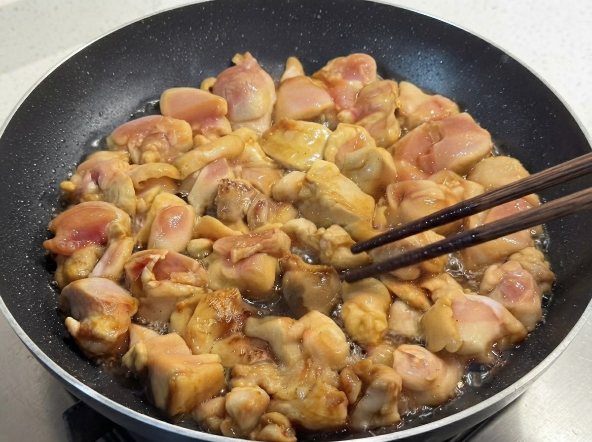 フライパンで炒められている下味付きの鶏肉を、木製の箸で丁寧に裏返している様子。