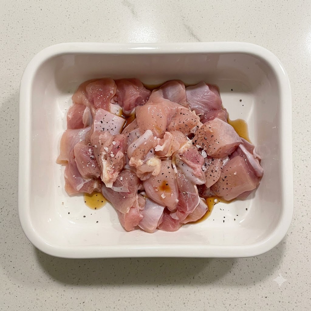 白い陶器の容器の中で、片栗粉、醤油、黒こしょうで下味をつけられた生鶏肉の切り身。