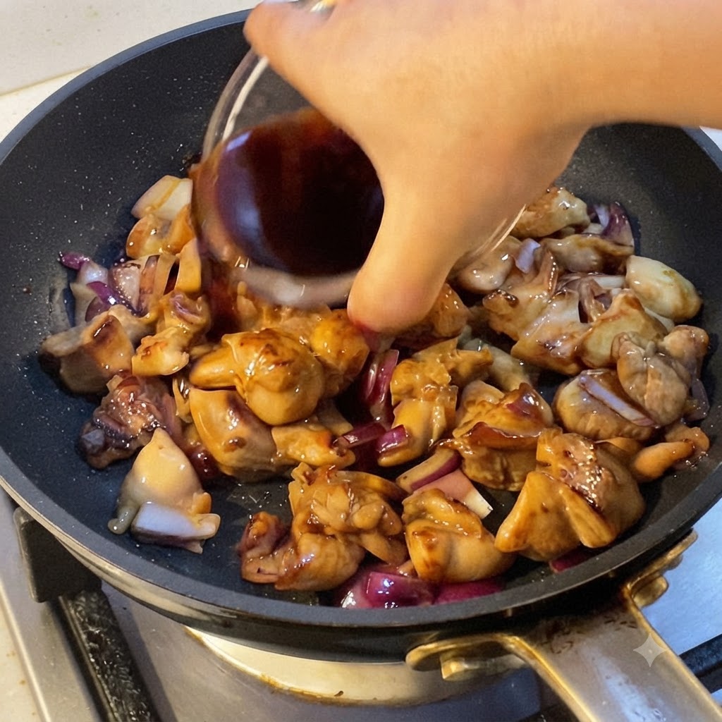 火の通った鶏肉と赤玉ねぎの入ったフライパンに、濃い色の調味液を注ぎ入れる手。