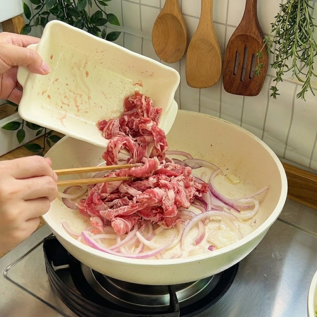 炒めた玉ねぎが入ったフライパンに、生の牛肉の薄切りを加えて炒めている様子。