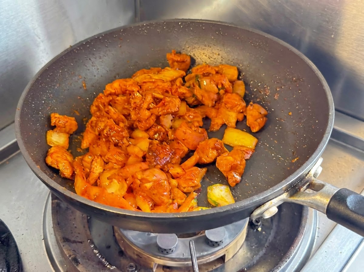 黒いノンスティックパンの中で、さいの目に切った鶏胸肉の玉ねぎと刻んだキムチが一緒に炒められている様子。