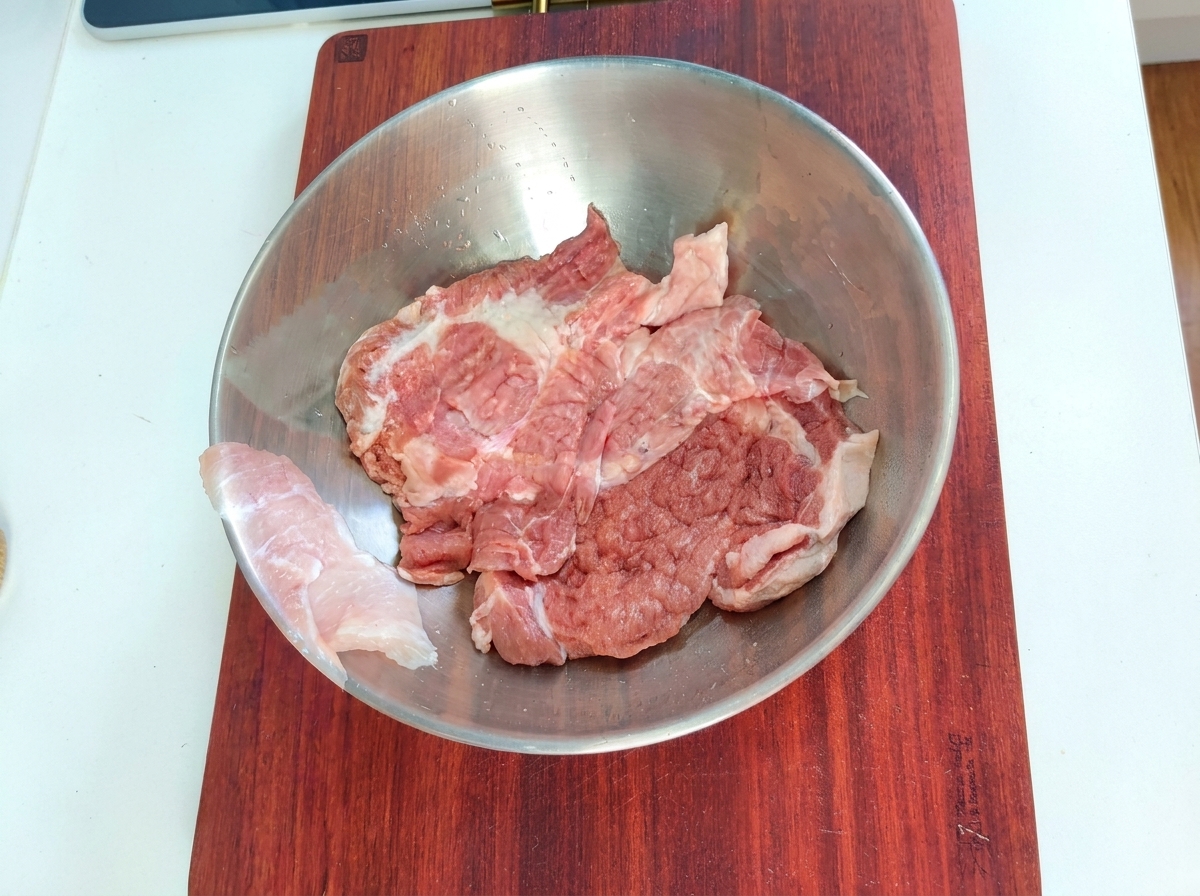 金属製のボウルに入った豚肉に、マリネ用の調味料が加えられている様子。