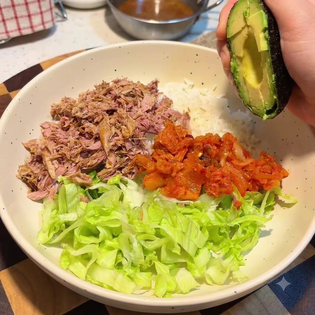 ご飯、レタス、ツナ、キムチが入った大きなボウルの上で、アボカドの半身を持っている手。