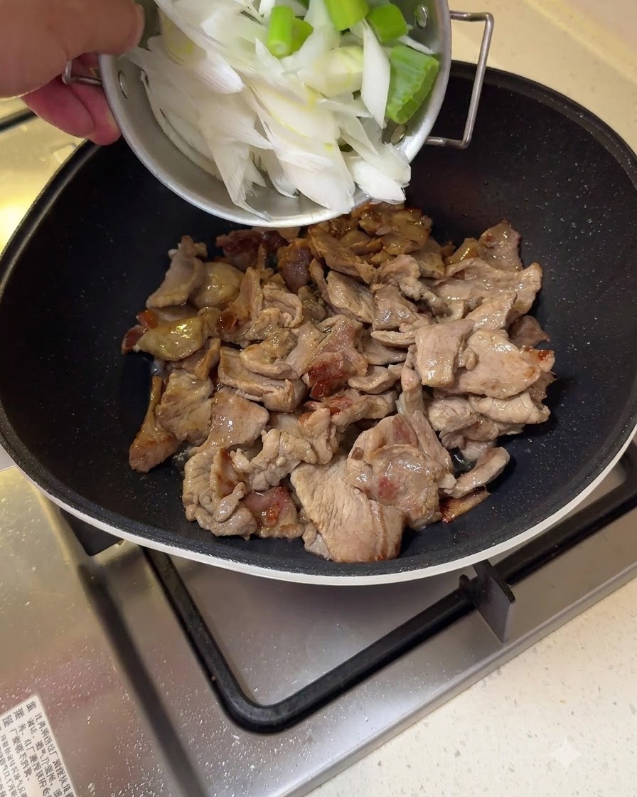 フライパンで炒めている豚肉に、大きく切った長ネギを加えている様子。
