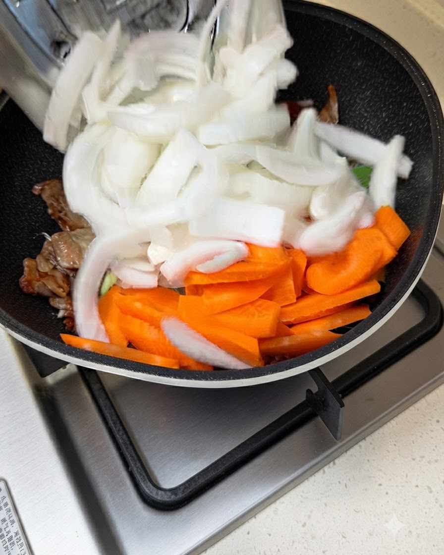 肉とソースが炒められているフライパンに、スライスした白玉ねぎとニンジンを入れている様子。