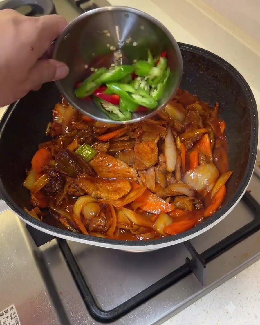 小さなボウルから緑と赤の唐辛子のスライスを、炒められた豚肉と野菜のフライパンに投入している様子。