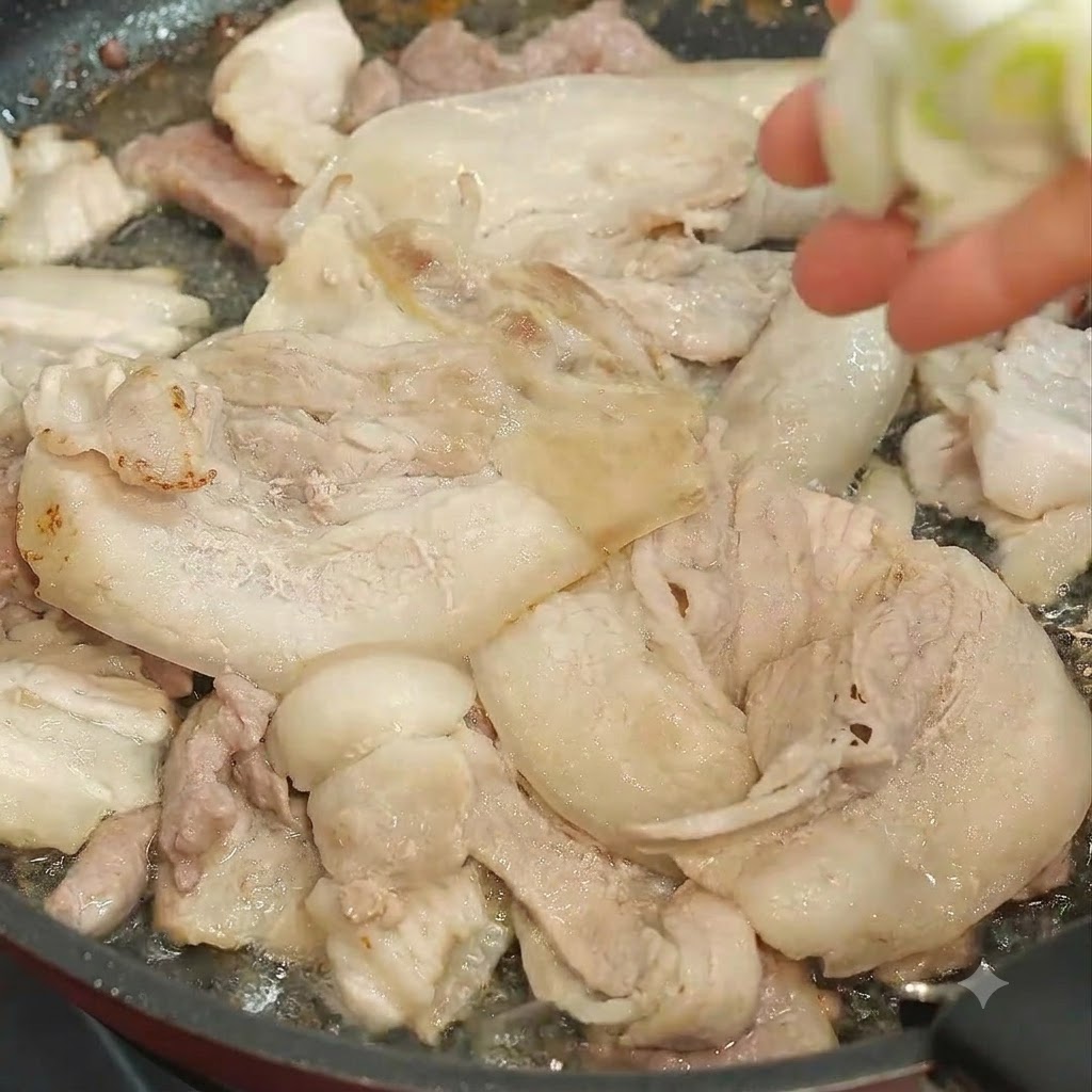 炒めている豚肉が入ったフライパンに、ざく切りにした青ねぎを手で投入している瞬間。