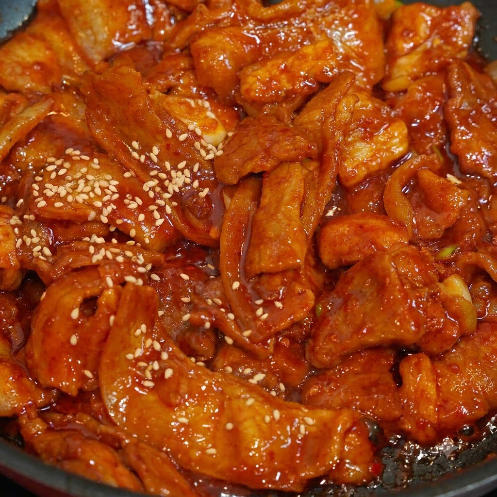 フライパンの中でタレに絡まった鮮やかな赤色のピリ辛豚肉に、白いりごまが振りかけられているクローズアップ。