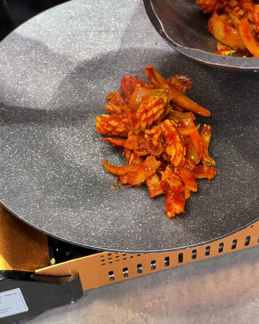 調理された赤いイカと豚肉の炒め物を、カセットコンロに設置したスキレットの中央に盛り付けている様子。