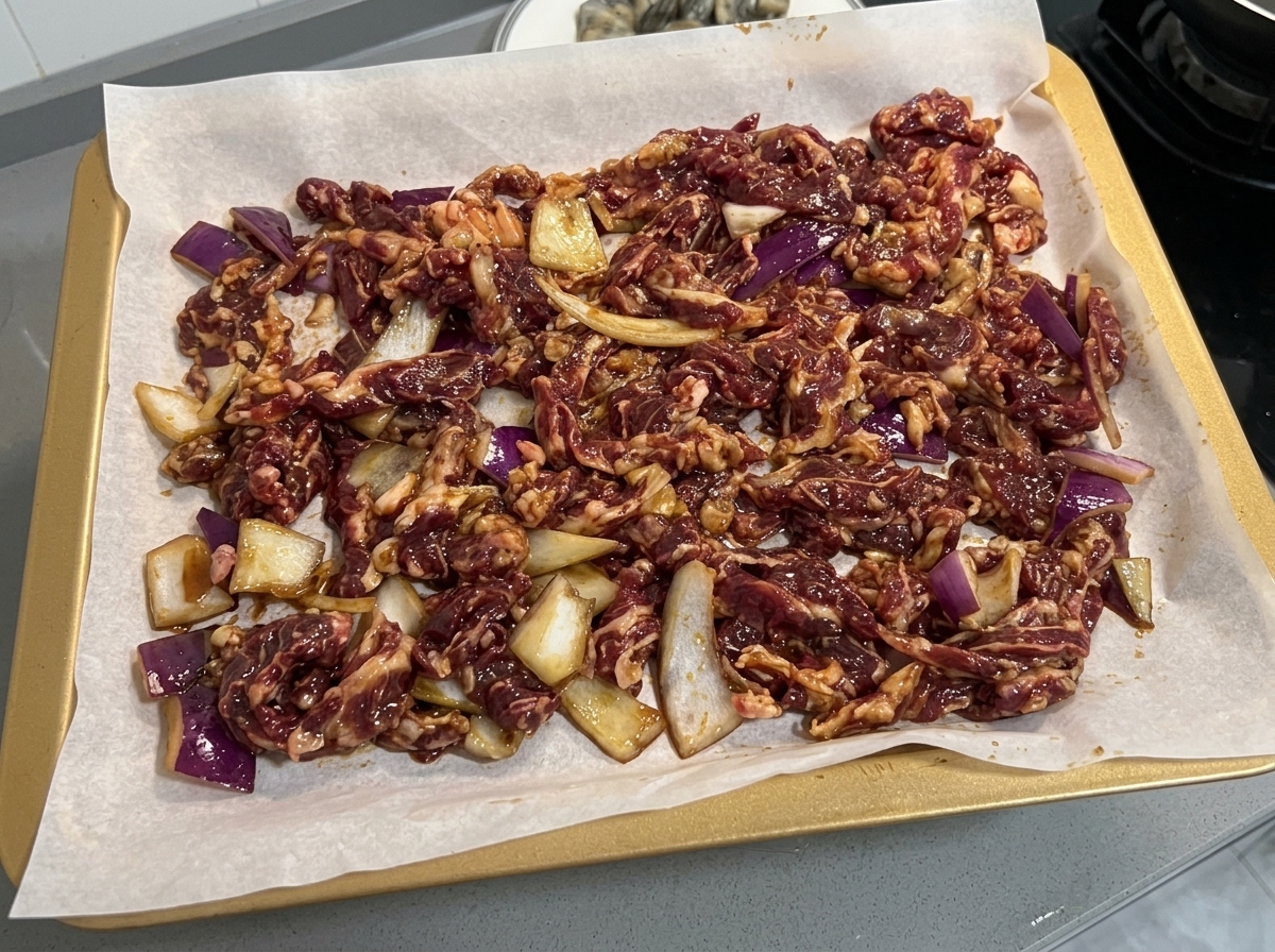 クッキングシートを敷いた天板に、下味をつけた牛肉と玉ねぎのスライスを均一に広げている様子。