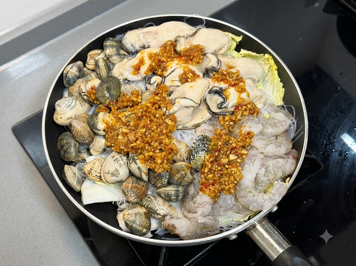 フライパンの中の生の状態のシーフードと麺類の上に、調理したガーリックとチリのソースをたっぷりと回しかけている様子。