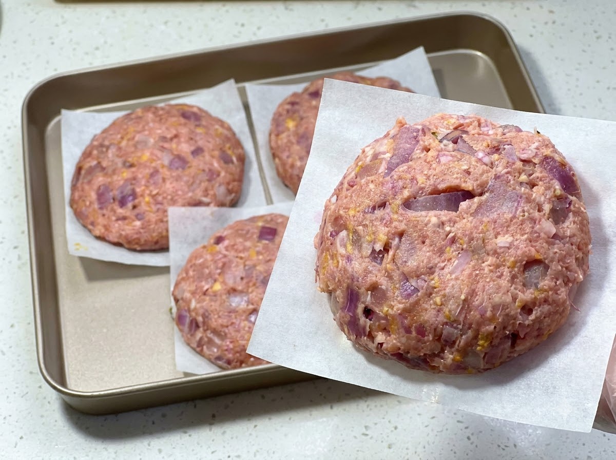 クッキングシートの上にのせられた生ハンバーグと、後ろのトレイに並べられた他のパティ。