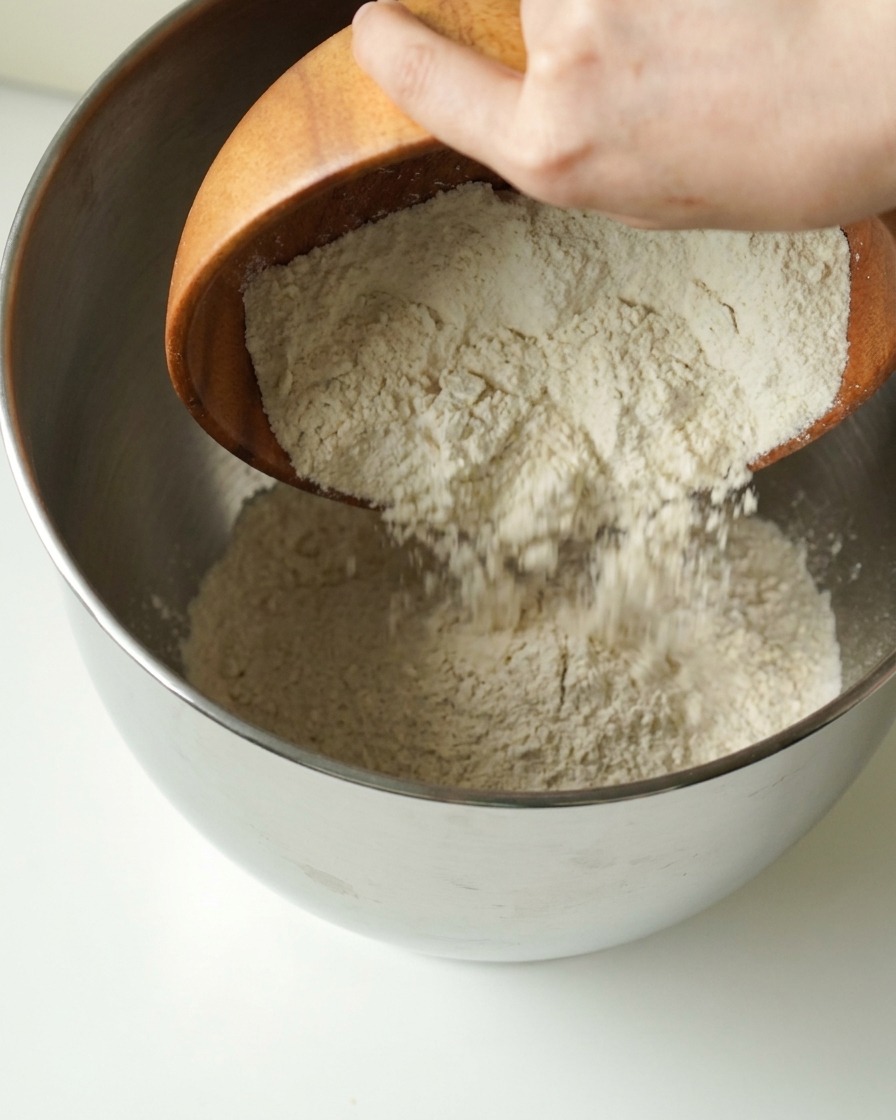 木のボウルから金属製のミキシングボウルに小麦粉が注がれている様子。