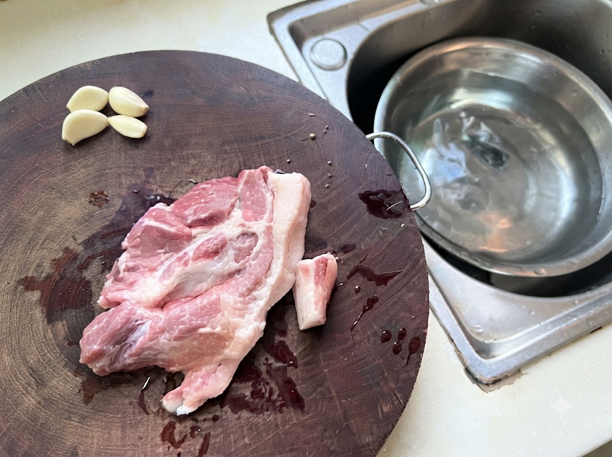 水が入った金属製ボウルの隣で、木製のまな板の上に広げられた豚ひき肉とニンニクの欠片。