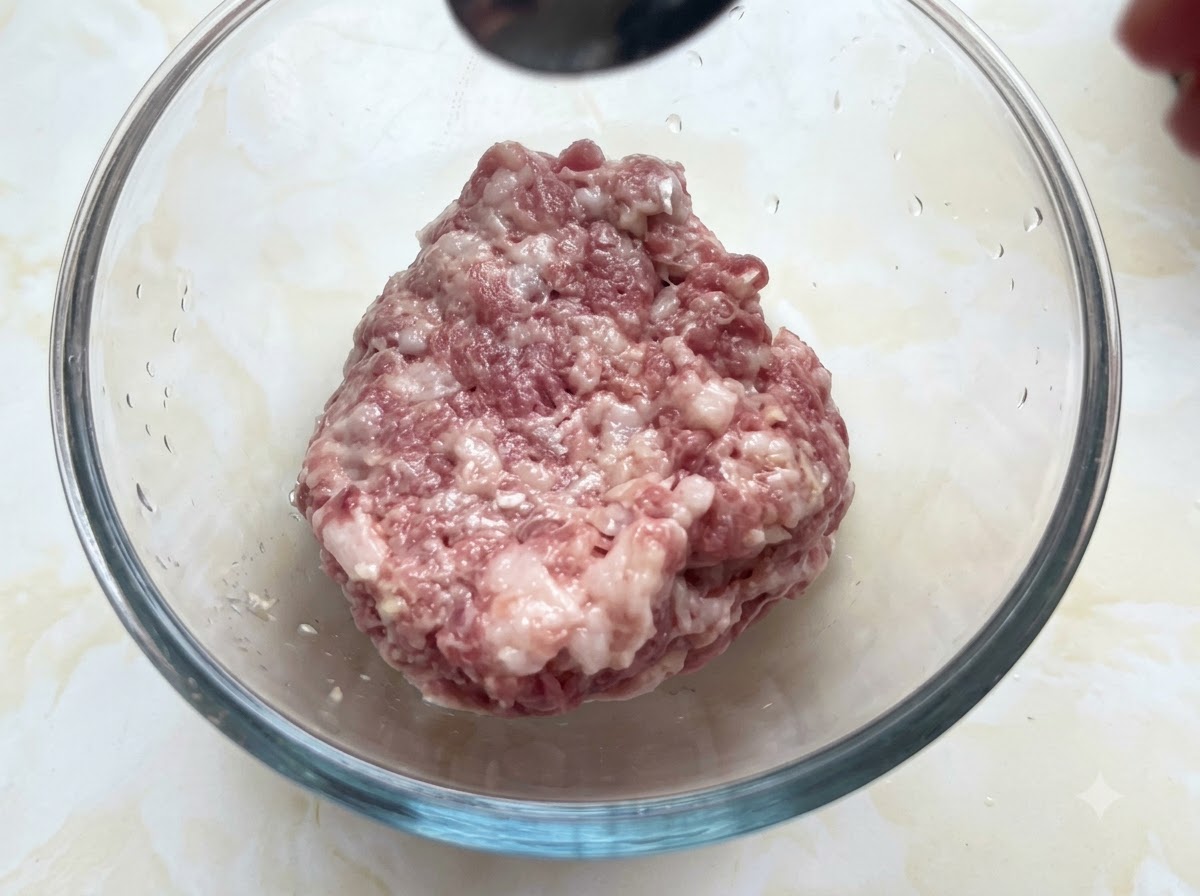 上からスプーンで調味料が加えられている、豚ひき肉が入ったガラス製ボウル。