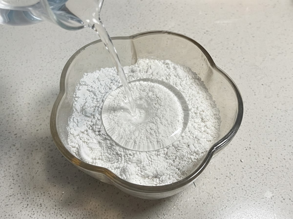 計量カップから、小麦粉と澱粉の入ったガラスボウルへ水が注がれている様子。