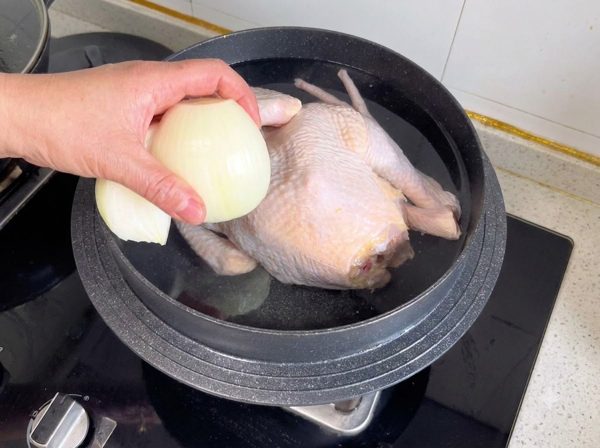 丸鶏と水が入った鍋に、半分に切った玉ねぎなどの香味野菜を入れている様子。