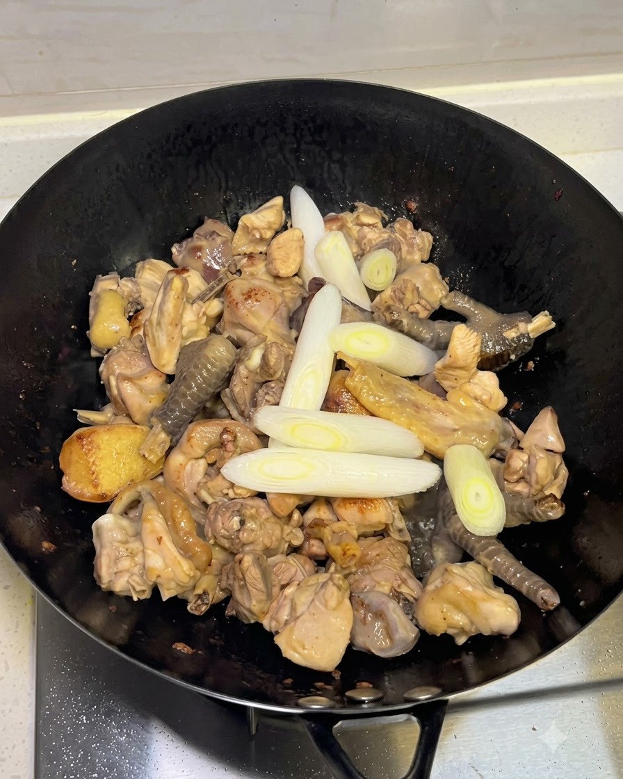 鶏肉の入った中華鍋に、ネギ、ニンニク、ホールスパイスを加えている様子。