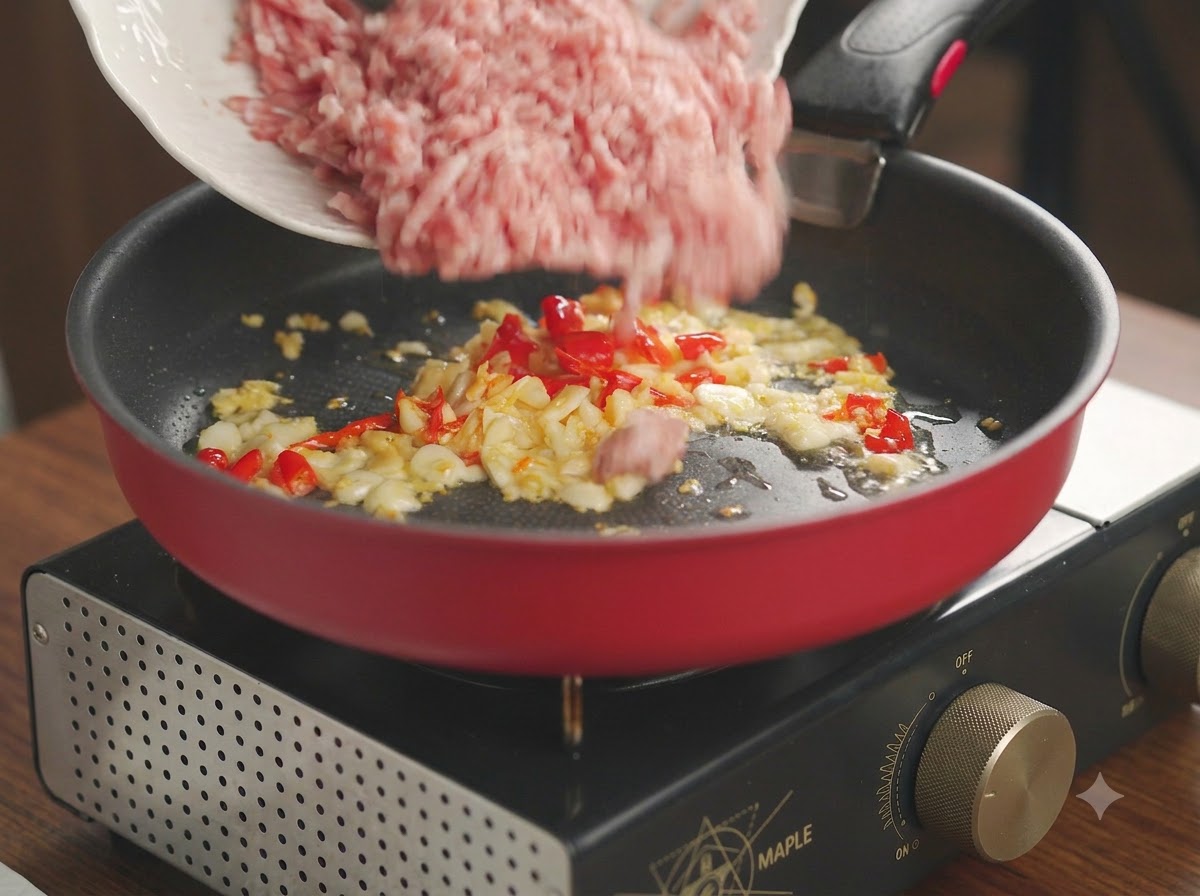 炒めたニンニクと唐辛子の入った赤いフライパンに、白い皿から投入される生の豚ひき肉。
