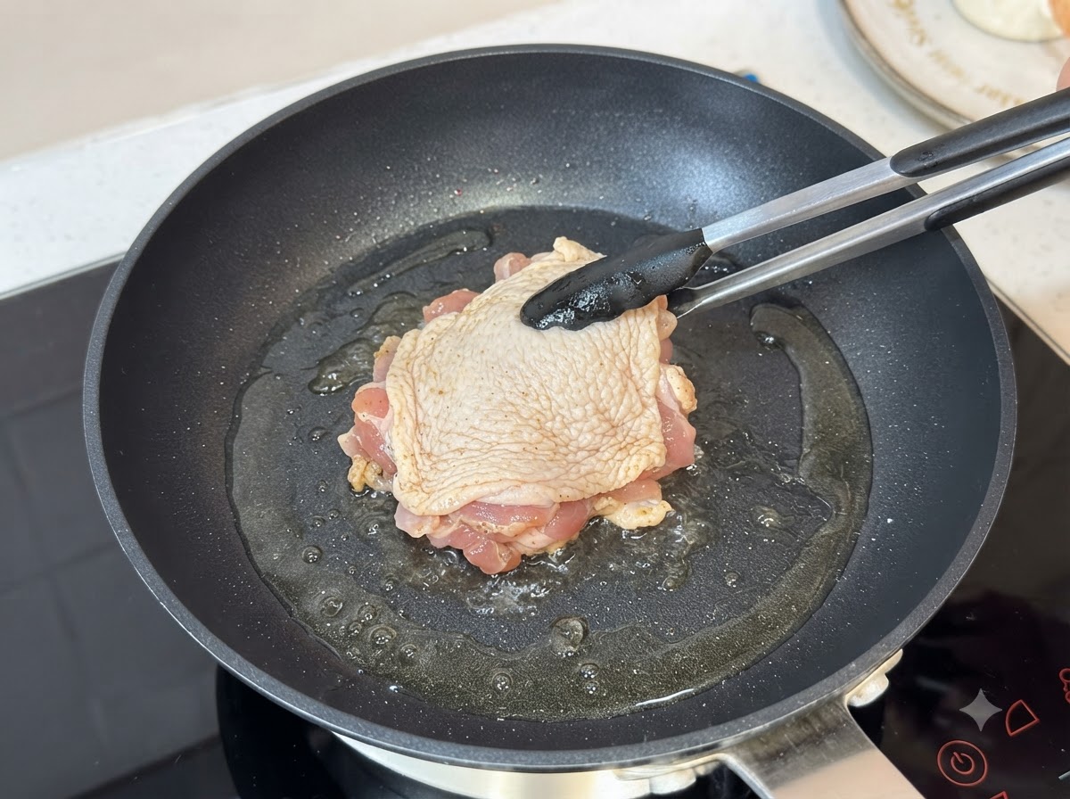熱した油を引いたフライパンで、トングを使い皮を下にして鶏もも肉を揚げ焼きにしている様子。