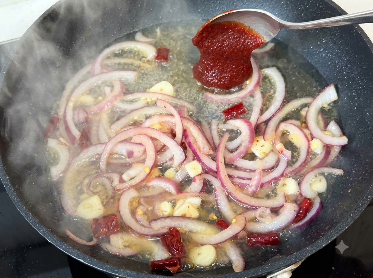 玉ねぎやスパイスが入った煮込み中のフライパンに、スプーン一杯のコチュジャンが加えられている様子。